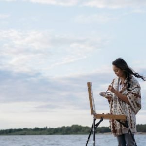 Pensive asian artist painting picture on canvas outdoor with clouds on background. Portrait of cheerful Korean woman painter holding paintbrush and palette, admire beautiful landscapes